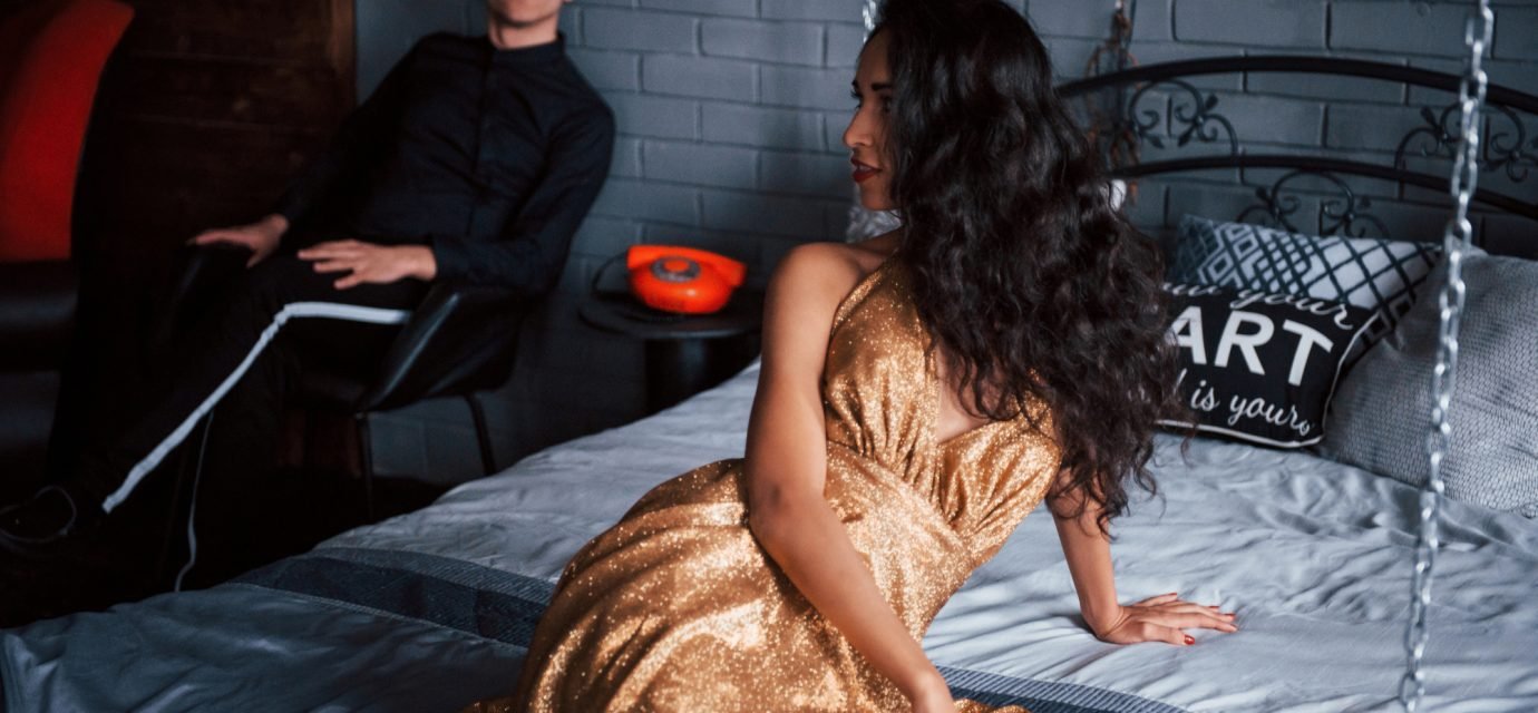 Swingers Club - Girl turned to look at her husband. Couple in classic wear sits in front of the beautiful luxury decorated bed