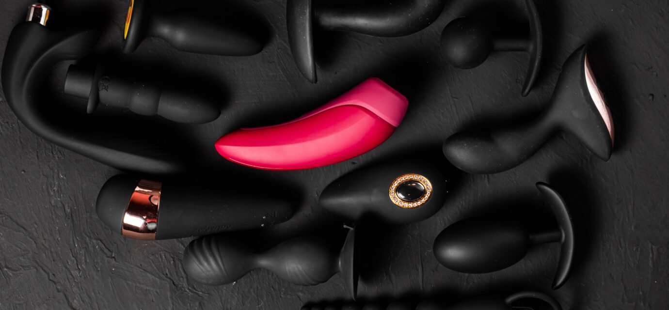 Erotic pleasure toys on a black background. Sex gadgets and masturbation devices. Pink dildo surrounded by black rubber and silicone sex toys: anal butt plugs, vibrators, vaginal balls. Top view.