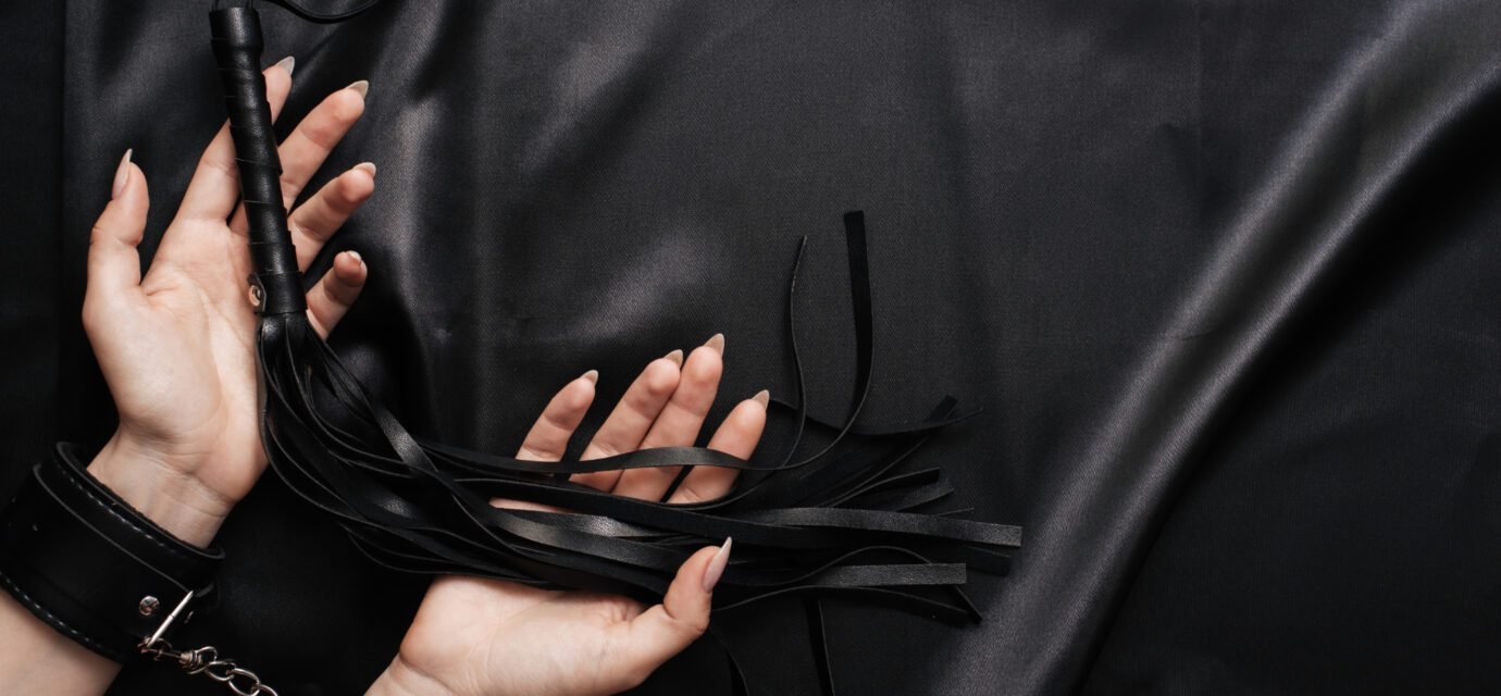 female hands in handcuffs on a dark silk sheet with a whip