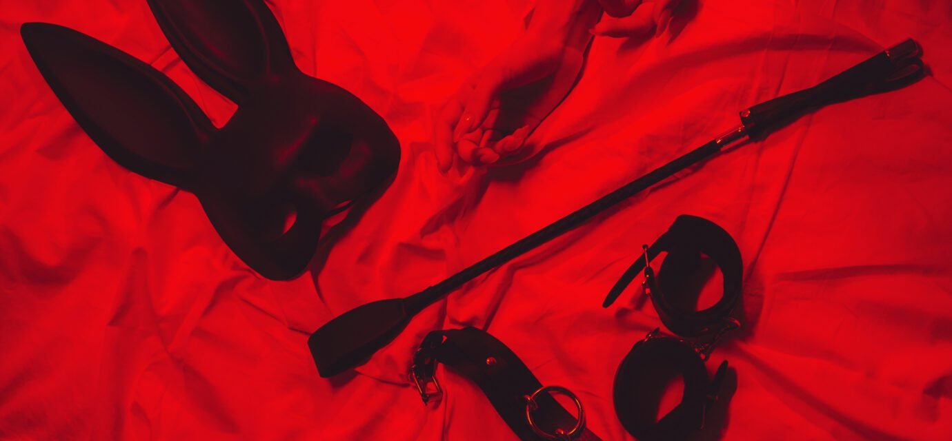 set of BDSM accessories with female hands is lying on the bed. Flogger whip, handcuffs, mask and choker for sex